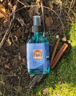 Aftershave Blue Wood Brodaty Ślązak na tle lasu, w otoczeniu zielonego jabłka, mchu i laski cynamonu – zdjęcie produktowe